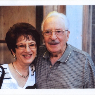 Colour photograph of an elderly couple smiling arm-in-arm at the camera. Both wear glasses, and the woman has dark brown short hair.