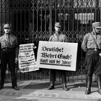 Black-and-white photograph of three men in military uniforms, standing in front of a closed storefront that has iron gates. Two of the hold up signs written in German. The other soldier is in discussion with a civilian.