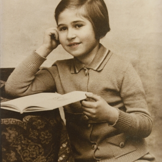 Photo de couleur sépia d'une jeune fille assise à une table sur laquelle il y a une nappe. La fille retient un livre ouvert sur la table d'une main et s'appuie sur l'autre d'une manière pensive. Un léger ton de rose a été ajouté à ses lèvres.