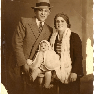 Sepia-tone photograph of a man and woman posing with a baby for a portrait. The man wears a hat and suit and tie, and the woman also wears a hat. The baby wears a white cape. The photograph’s edges have some wear and tear.