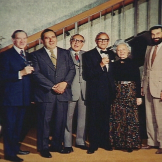 Photo en couleur d'un groupe de huit personnes se tenant debout en ligne et souriant à la caméra. Les sept hommes portent des complets et la femme du groupe porte un robe longue.
