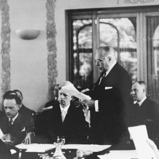 Black-and-white photograph of a man standing among a room of men who are seated at desks. The man standing up reads aloud from the paper he is holding, while wearing glasses and a business suit.