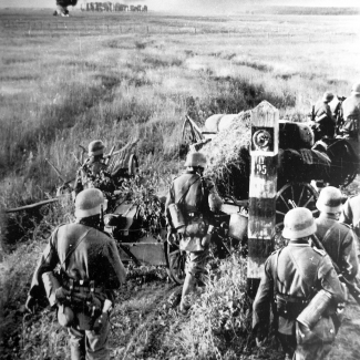 Black-and-white photograph of a military troop of about half a dozen men walking into a field. There appears to be a large fire with a cloud of dark smoke in the far distance.