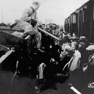 Photo en noir et blanc d'un homme assis sur le bord d'une voiture de train, tenant une canne, et  regardant une plateforme remplie de gens.