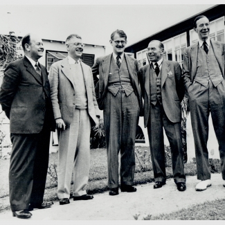 Black-and-white photograph of five men, wearing suits, standing outside in a row and smiling or laughing.