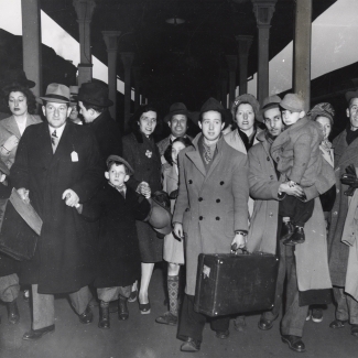 Black-and-white photograph a group of about 16 people walking towards the camera on a train platform. They are wearing jackets and some carrying suitcases. There are four young children in the group.