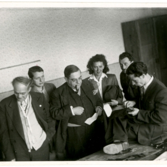 Black-and-white photograph of a group of six people, five men and one woman, standing in a room. One man is smoking a pipe and observing another man sitting on a table, hand sewing fabric.