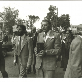 Black-and-white photograph of four men leading a march outdoors. The man in the centre is carrying a rectangular box. A group of people follows them in the background.