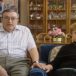 Photo en couleur d'un homme et d'une femme âgés, se tenant la main alors qu'ils sont assis ensemble à l'intérieur. La femme est assise sur un fauteuil bleu. Il y a un vaisselier en arrière-plan dans lequel il y a des plats et de la porcelaine en arrière-plan.