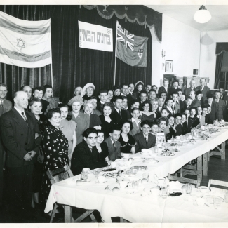 Photo en noir et blanc d'un grand groupe d'environ 70 personnes souriant pour une photo de groupe dans une salle. Le groupe est installé derrière une grande table sur laquelle sont disposés des plats et de la nourriture, et deux drapeaux sont accrochés sur des rideaux en arrière-plan.