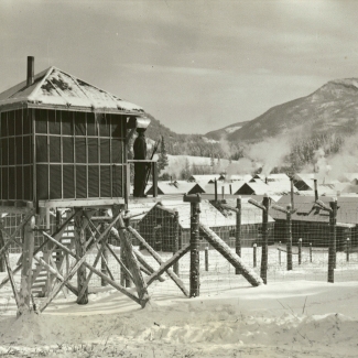 Black-and-white photograph of a guard standing at attention in his barracks, surrounded by barbed wire. Rows of buildings are in the background, with snow-covered hills in the distance. There is snow on the ground.