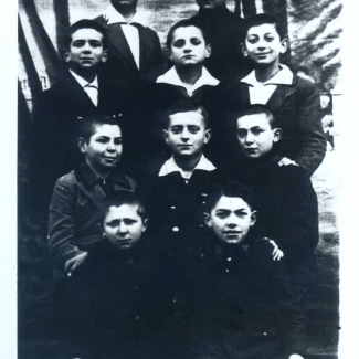 Black-and-white photograph of a group of ten young boys, posing in four rows of two or three. The boys look at the camera, most of them wearing collared shirts under jackets.