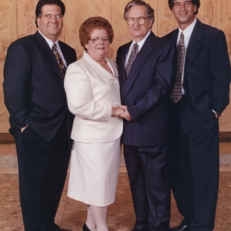 Photo en couleur prise en studio d'un groupe de quatre adultes se tenant debout ensemble et souriant à la caméra. Le couple au centre est plus âgé et la femme porte un veston et une jupe blancs. Les trois hommes portent des complets.