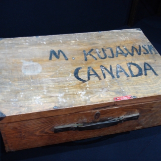 Photo en couleur d'une valise en bois de forme rectangulaire avec les mots “M KUJAWSK/CANADA” peints sur le dessus de la valise.