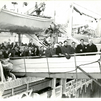 Black-and-white photograph of a group of 15 people, wearing coats and hats, smiling and waving at the camera from the gangway of a ship. Several more people wait to disembark the ship. A lifeboat is suspended in the top-left corner with ice frozen to its sides.