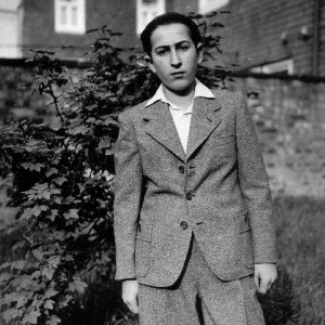Photo en noir et blanc d'un jeune homme debout à l'extérieur dans ce qui semble être une cours arrière avec une clôture et un arbuste derrière lui. Il porte un complet et une chemise à col, ses cheveux sont coiffés vers l'arrière.