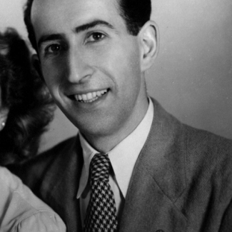 Black-and-white studio photograph of a man sitting and smiling at the camera. The man wears a suit and has dark brown hair.”