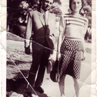 Photo en noir et blanc d'un homme et d'une femme debout sur une allée pavée à l'extérieur. Il y a des arbres en arrière-pplan. L'homme porte un complet et la femme porte une blouse et une jupe à pois.