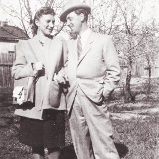 Black-and-white photograph of a man and woman, standing together, arm-in-arm, in a field outdoors with trees in the background. The couple smiles at one another and the man wears a hat and suit.