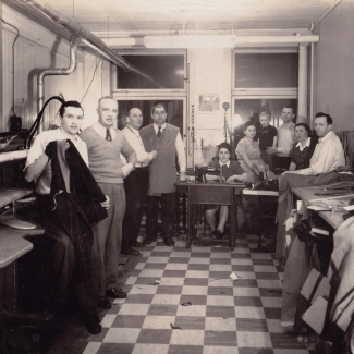 Photo en noir et blanc d'un groupe d'environ dix personnes debout ou assises ensemble dans une salle dans laquelle il y a deux fenêtres au fond. Le groupe regarde la caméra. Il y a des piles de tissus et de vêtements à droite et le plancher est à motif de damiers.