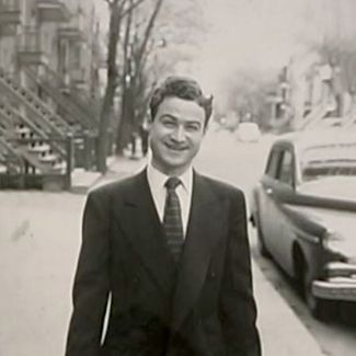 Black-and-white photograph of a man standing outside on a sidewalk, wearing a suit and smiling at the camera. The street behind him has a vintage car parked on the right, as well as trees, balconies, and stairwells leading up to apartments.