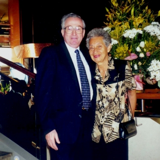 Colour photograph of an elderly couple standing and smiling together, arm-in-arm. The man wears a suit, and the woman wears a printed blouse. They stand in front of a large arrangement of flowers.