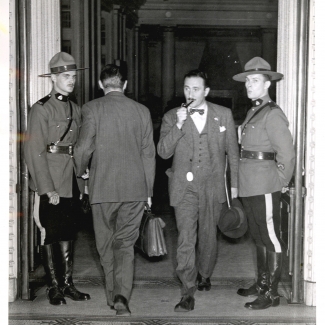 Black-and-white photograph of four men in a large doorway. One man, second from the right, is exiting the room with a briefcase and pipe in his mouth. The man to his left is entering the room. The two men on either side are dressed in RCMP uniforms and look towards the camera.