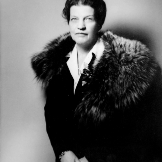 Black-and-white studio portrait photograph of a woman, pictured from the waist up, wearing a dark jacket with a fur stole. She looks directly at the camera.