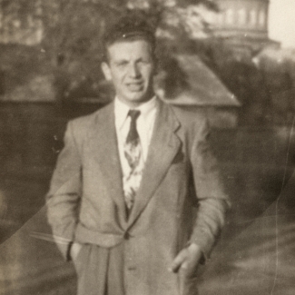 Black-and-white photograph of a man wearing a suit and tie, standing outdoors on a field. A building tower is visible in the background.