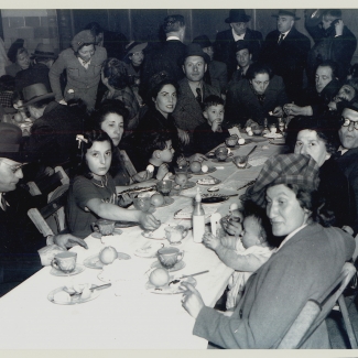 Black-and-white photograph of a large group of people sitting together at a long table, looking at the camera. The table is full of dishes and food. There are several more people in the room behind the table.