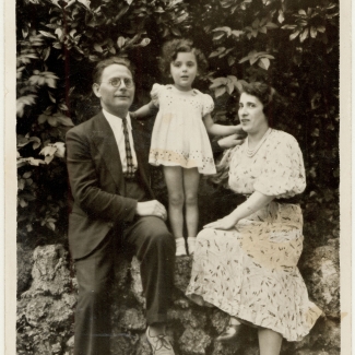 Black-and-white photograph of a man and woman sitting outdoors on a rock wall, with a young girl standing between on the wall between them, leaning on their shoulders for support. The woman and her daughter wear dresses, and the man is in a suit.