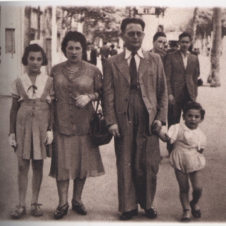 Sepia-toned photograph of a man and woman, holding the hands of their two children, standing on either side of them. The family of four is outside on a street or walkway, with people walking in the background.