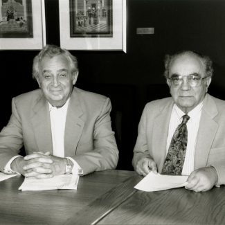 Photo en noir et blanc de deux hommes âgés assis ensemble à une table avec des papiers devant eux. Les hommes portent des vestons, l'un d'eux porte une cravate et il y a trois œuvres d'art encadrées et accrochées derrière eux.