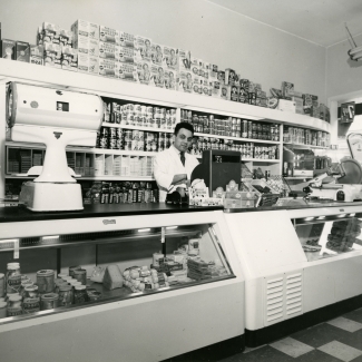 Black-and-white photograph of a man standing behind the cash register of  a large grocery store. He stands in front of a shelf full of food products, and there is a large display food counter in front of him.