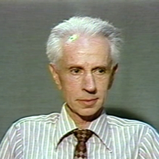 Colour photograph of a close-up shot of an elderly man with white hair positioned against a grey background, looking towards the right of the camera. He wears a white shirt with light blue stripes and a brown checkered tie.