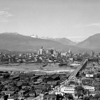 Black-and-white photograph of a bird's eye view of a city. Several buildings are pictured on either side of a river. A bridge connecting the two banks is pictured on the right-hand side of the photo. Several mountain peaks are visible in the background.