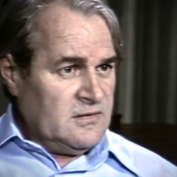Screenshot of Holocaust survivor Aba Beer video testimony. He is sitting in front of grey curtains, and looking to the right of the camera. The camera shows his face and shoulders.
