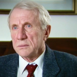 Capture d'écran du témoignage vidéo du survivant de l’Holocauste Elliott Zuckier, assis dans son salon, et regardant à la gauche de la caméra. Son visage et ses épaules sont visibles à la caméra.