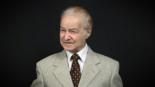 Capture d'écran de Leon Hirsch, survivant de l'Holocauste, durant l'enregistrement de son témoignage vidéo. Il est assis devant un fond gris et regarde à la gauche de la caméra. Son visage et ses épaules sont visibles à la caméra.